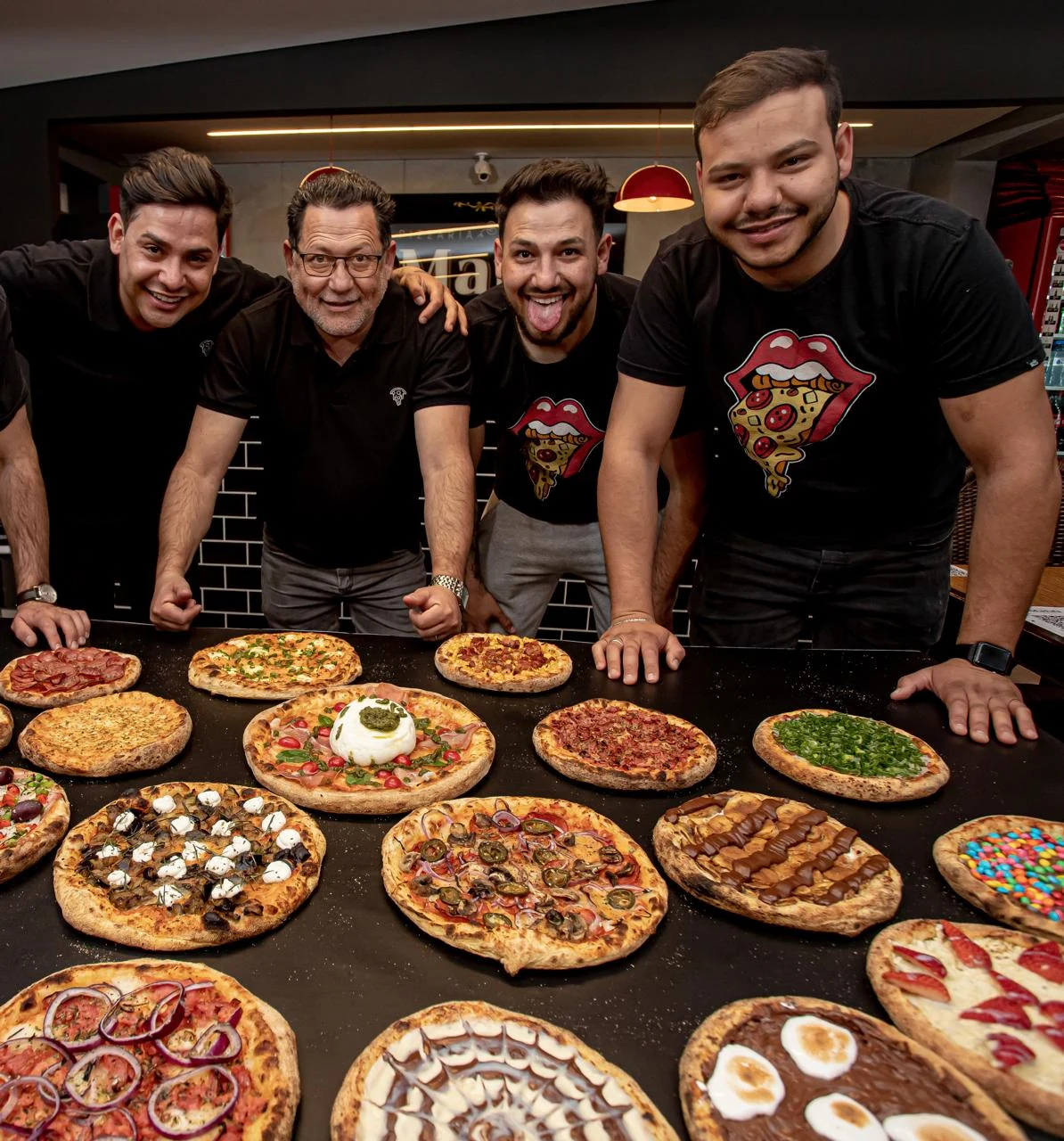 Família Maia reunida na pizzaria, atrás de uma bancada com pizzas.